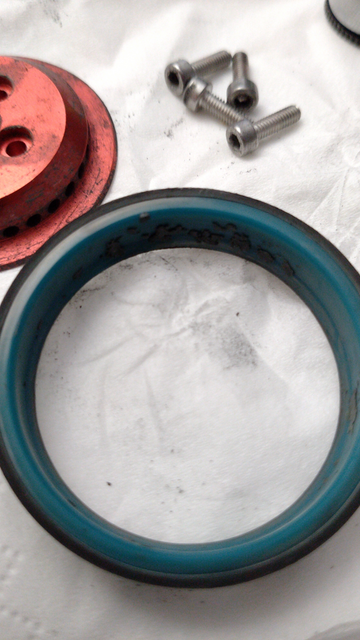 Ein dunkeltürkisfarbener Ring mit ein wenig dunklem Dreck drin liegt auf einer hellen Oberfläche. Darüber ein dreckiges, rot eloxiertes, rundes, deckelförmiges Metallteil mit Belüftungs- und Schraubenlöcher sowie vier passende Schrauben.