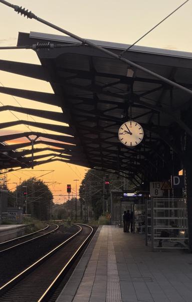 Ein ziemlich leerer Bahnsteig im Sonnenuntergang. Die Analoge Uhr zeigt kurz vor 10. die Signale stehen alle of rot. Die Schienen reflektieren in der Sonne.