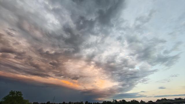 Links ein dunkelgraues, dichtes Wolkenband, rechts blauer Himmel, in der Mitte mausgraue Schäfchenwolken und ein schmales Band von in kräftigem Orange leuchtenden Wolken.